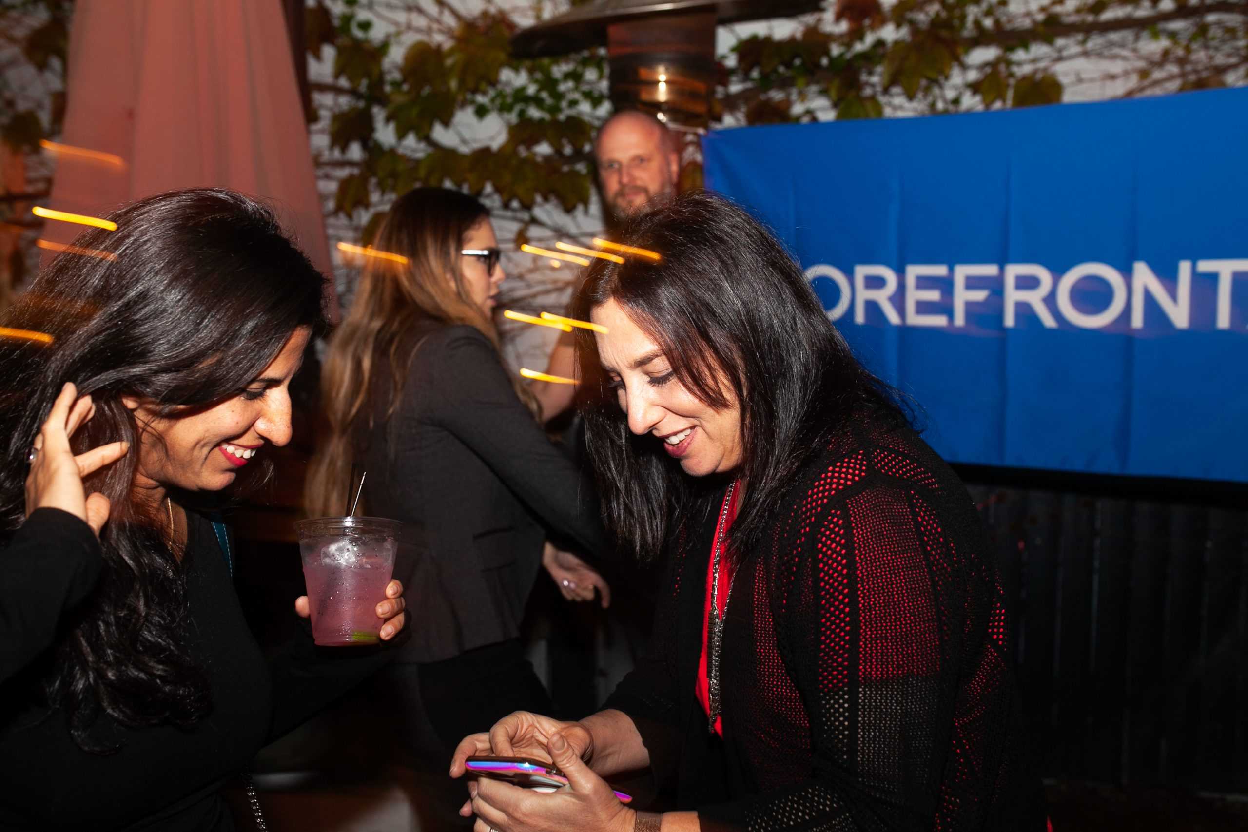 Two women smiling and laughing at an event, one is holding a drink and the other is looking at the screen of a phone.