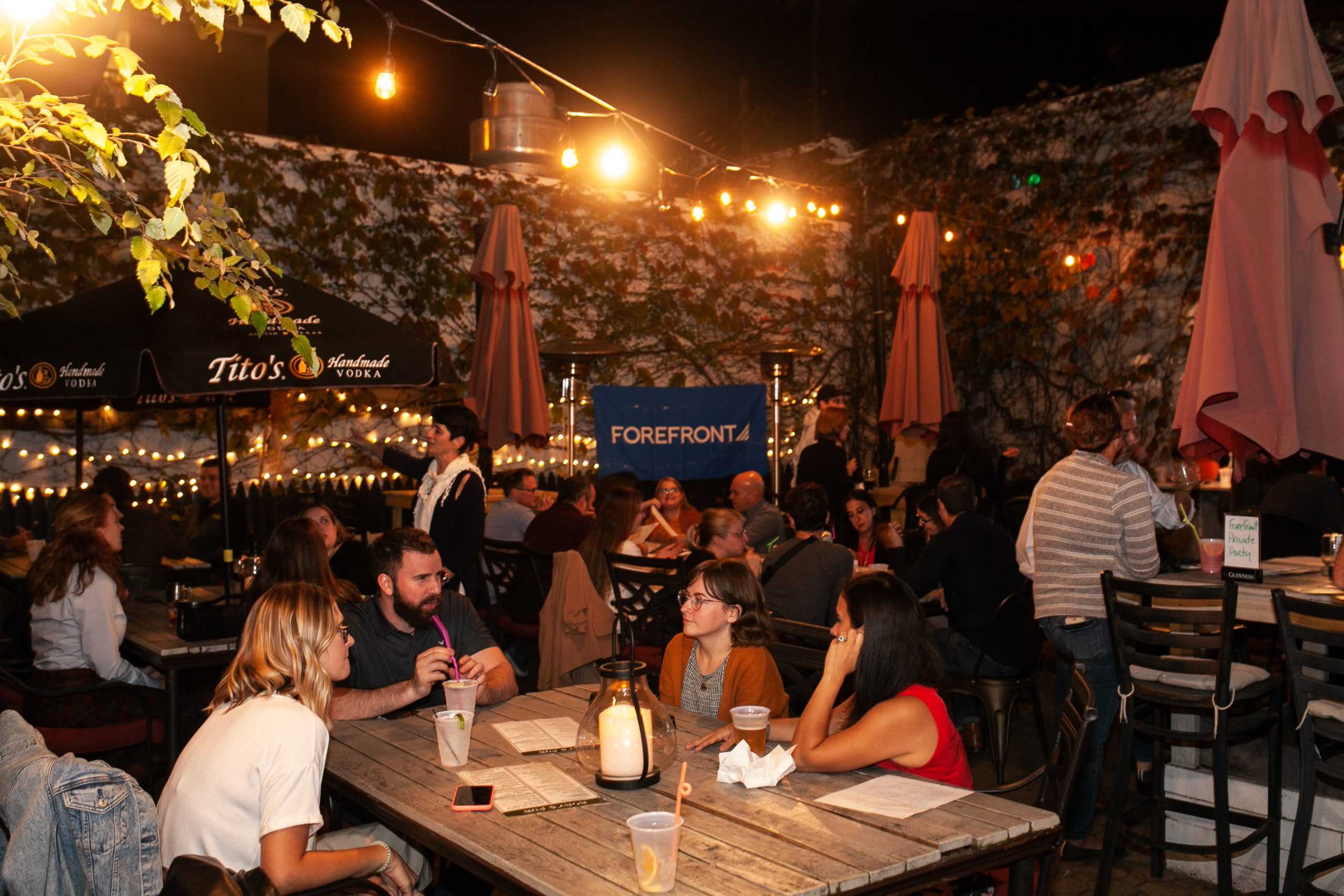 A group of 4 people sitting at a wooden table in a busy outdoor restaurant patio. There is a blue banner with the text "ForeFront" in the background.