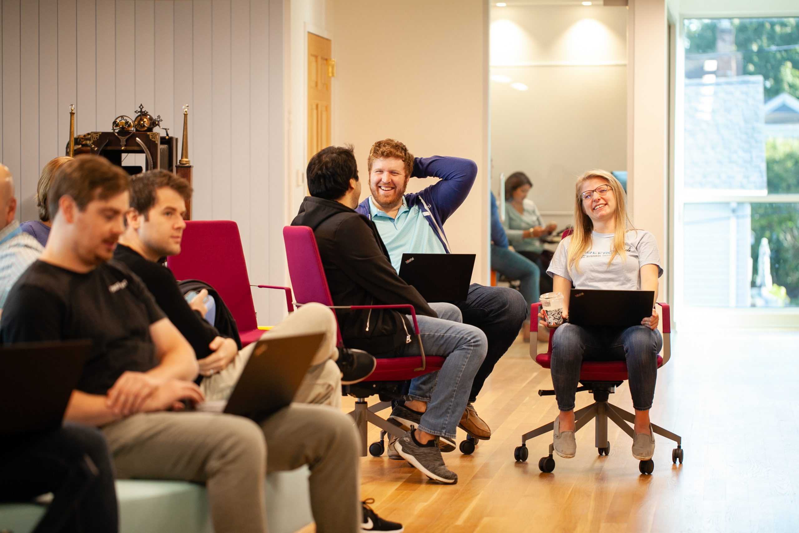 Five people sitting in a room working on laptops. Some are listening intently, others are laughing in conversation.