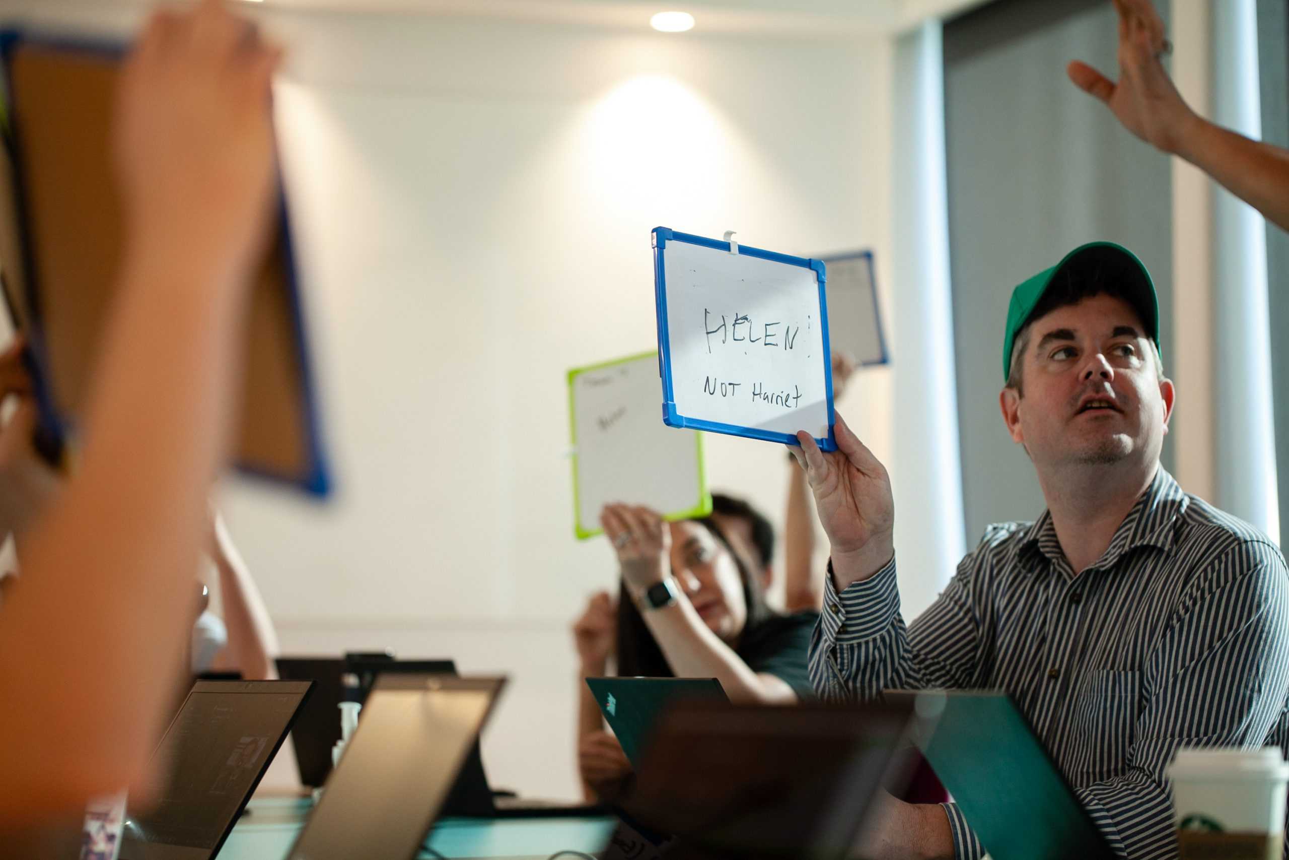 A man in a green cap holding up a small dry erase board with written words on it.