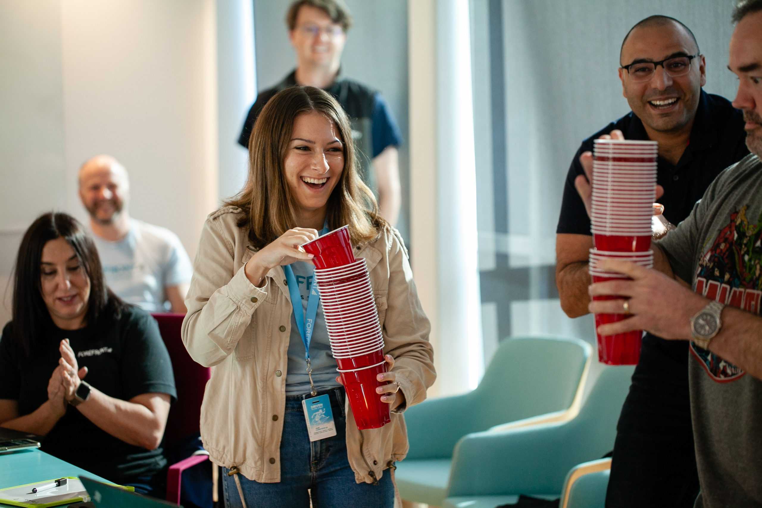 A woman smiling while carrying a tall stack of cups in a room full of people.