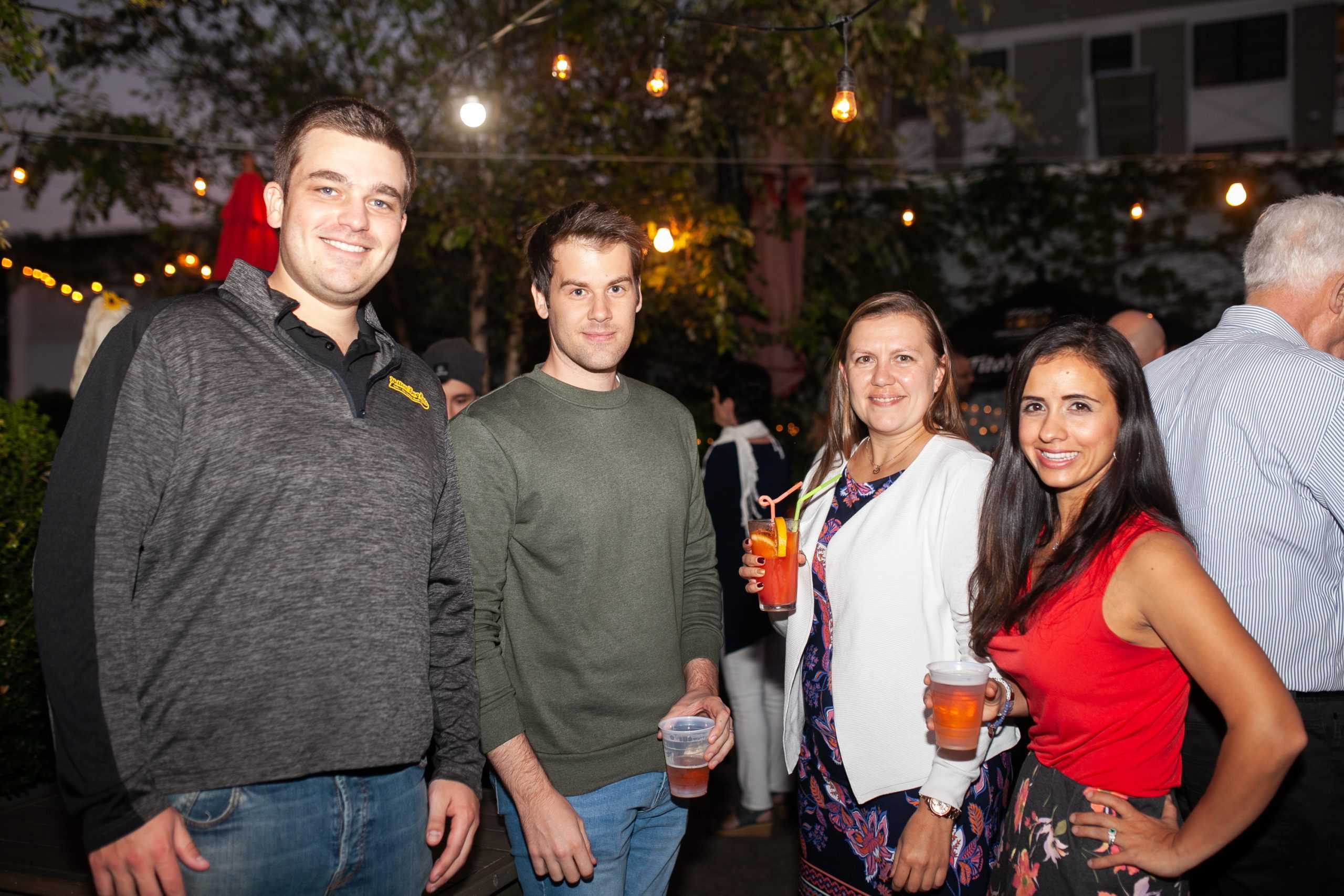 A group of four people at an event, three are holding drinks. Everyone is looking at the camera and smiling.