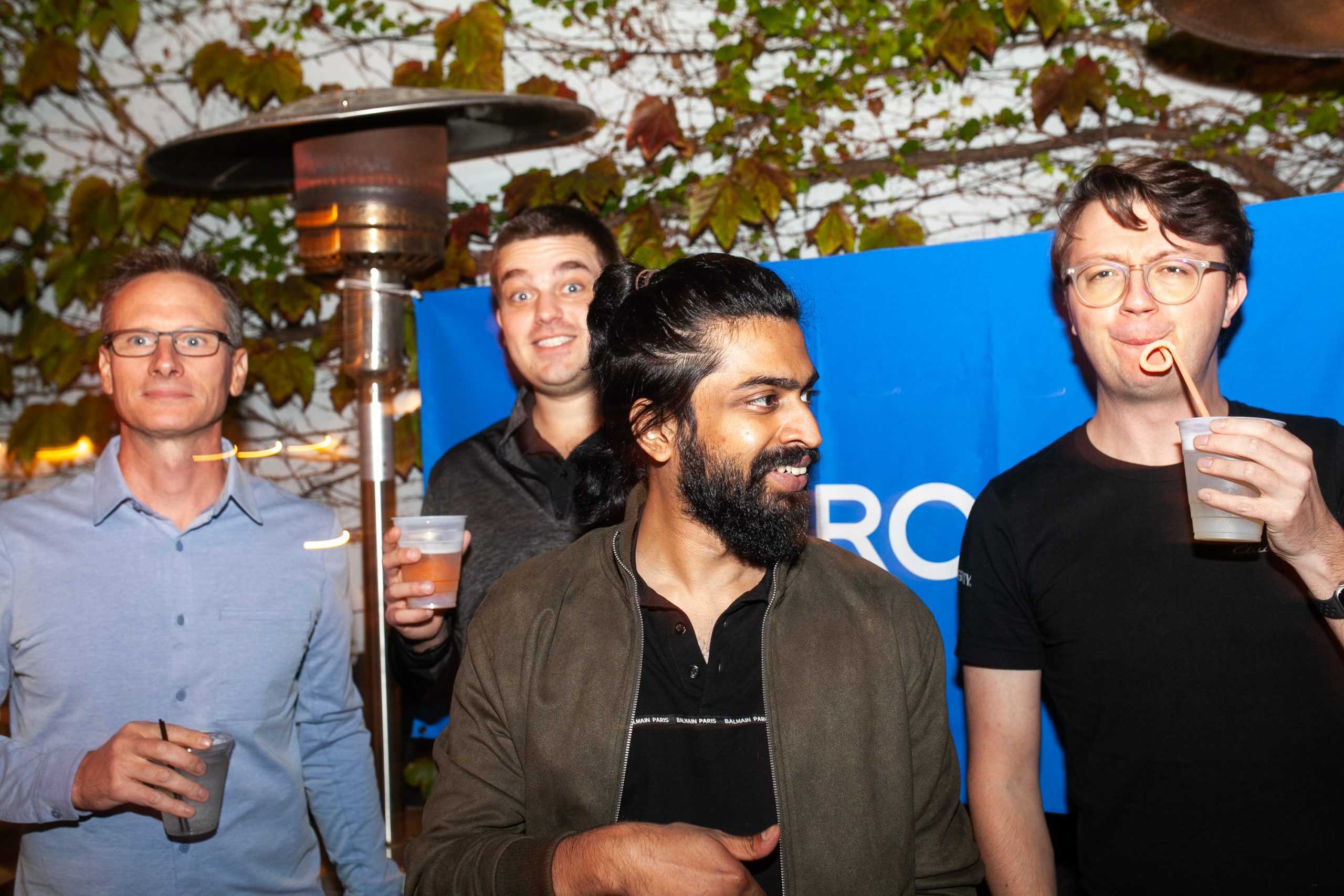 A group of four men are enjoying an outdoor gathering in front of a backdrop covered with green ivy and a blue banner. They are holding drinks and appear to be in a relaxed, festive mood. The man on the left wears glasses and a light blue shirt, while the man next to him in a dark shirt has a surprised expression. The man with a beard and dark hair pulled back is in the center, looking to his right. The man on the right is sipping from a cup with a curly straw, wearing glasses and a black shirt. A tall patio heater is also visible behind them, indicating it's an evening event.
