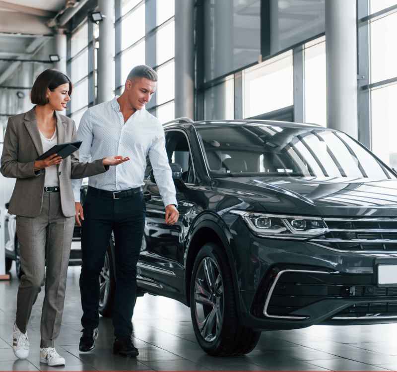 A woman and a man are walking beside a sleek, dark-colored SUV in a brightly lit car showroom. The woman, holding a tablet, is explaining something to the man, who is looking at the vehicle. The large windows and modern design of the showroom provide a professional and inviting atmosphere.