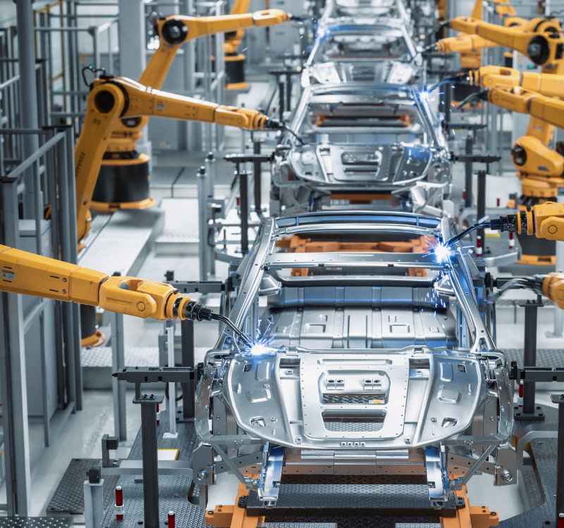 An image of a series of partially assembled cars being worked on in a manufacturing assembly line. Yellow robotic arms appear to be welding the unfinished frames of each car.