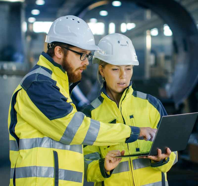 A man and a woman wearing yellow reflective jackets and white safety helmets. The woman is holding a laptop, and they are both looking at the screen intently.