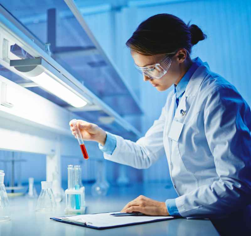 A woman in a brightly lit lab, wearing a white lab coat and holding a vial containing a red liquid. There is a clipboard on the table and a pen for note taking.