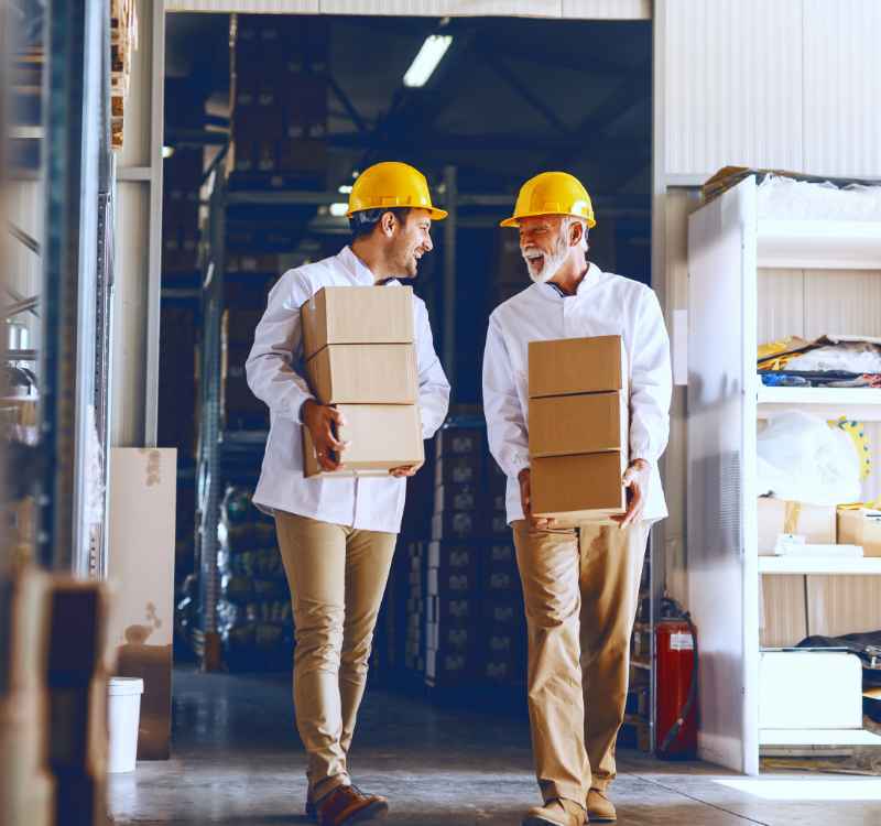Two men in a warehouse walking, talking, and smiling. Each man is holding three small cardboard boxes. They are wearing khaki pants, white long sleeve shirts, and yellow safety helmets.