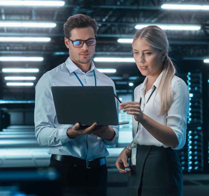 Two professionals, a man and a woman, in business attire standing in front of a laptop, engaged in a discussion.