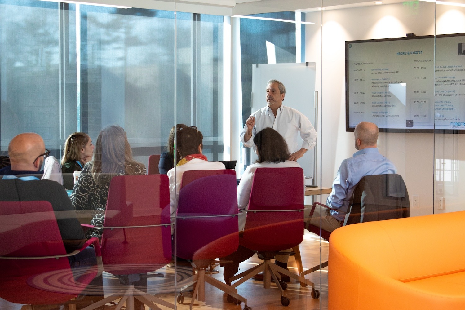 Home – WhyForefront_image A group of people is seated in a modern conference room with glass walls. They are facing a man standing at the front of the room who is giving a presentation. The presenter is dressed in a white shirt and is gesturing as he speaks. The attendees are seated in colorful chairs, an orange couch is partially visible in the foreground. The room is well-lit with natural light filtering through the window shades.