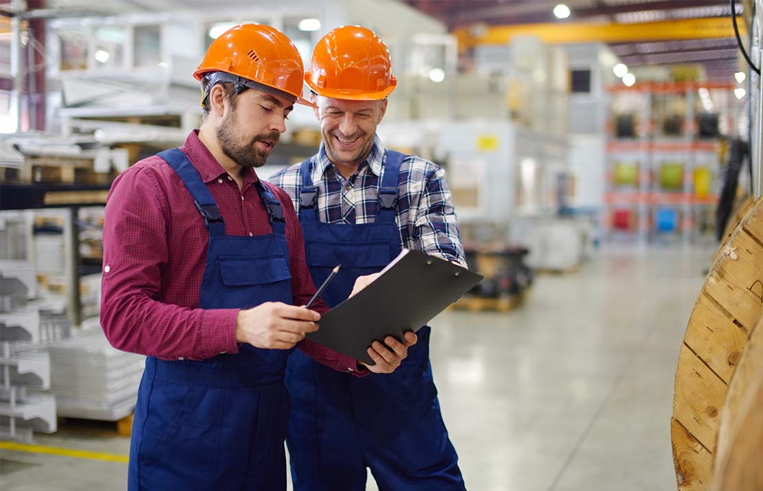Manufacturing-image Two workers wearing orange hard hats and blue overalls are standing in a spacious, well-lit industrial warehouse. They are smiling and engaged in a discussion while looking at a clipboard that one of them is holding. The background features shelves stocked with materials and industrial equipment.
