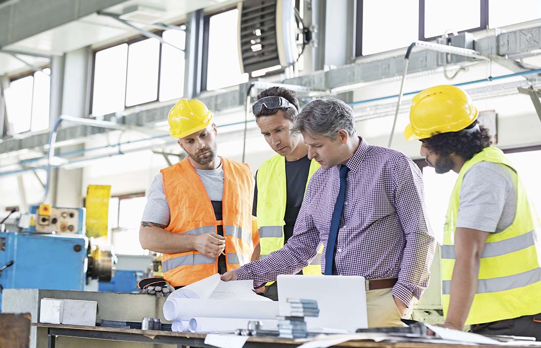 Manufacturing-image2 Four men in a warehouse environment looking at blueprints. Three of the men are wearing reflective safety vests and other safety gear like helmets and protective glasses, one is wearing a button up shirt and tie.