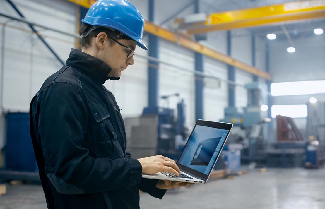 Manufacturing-image4 A man in a warehouse wearing a blue safety helmet holding a laptop.