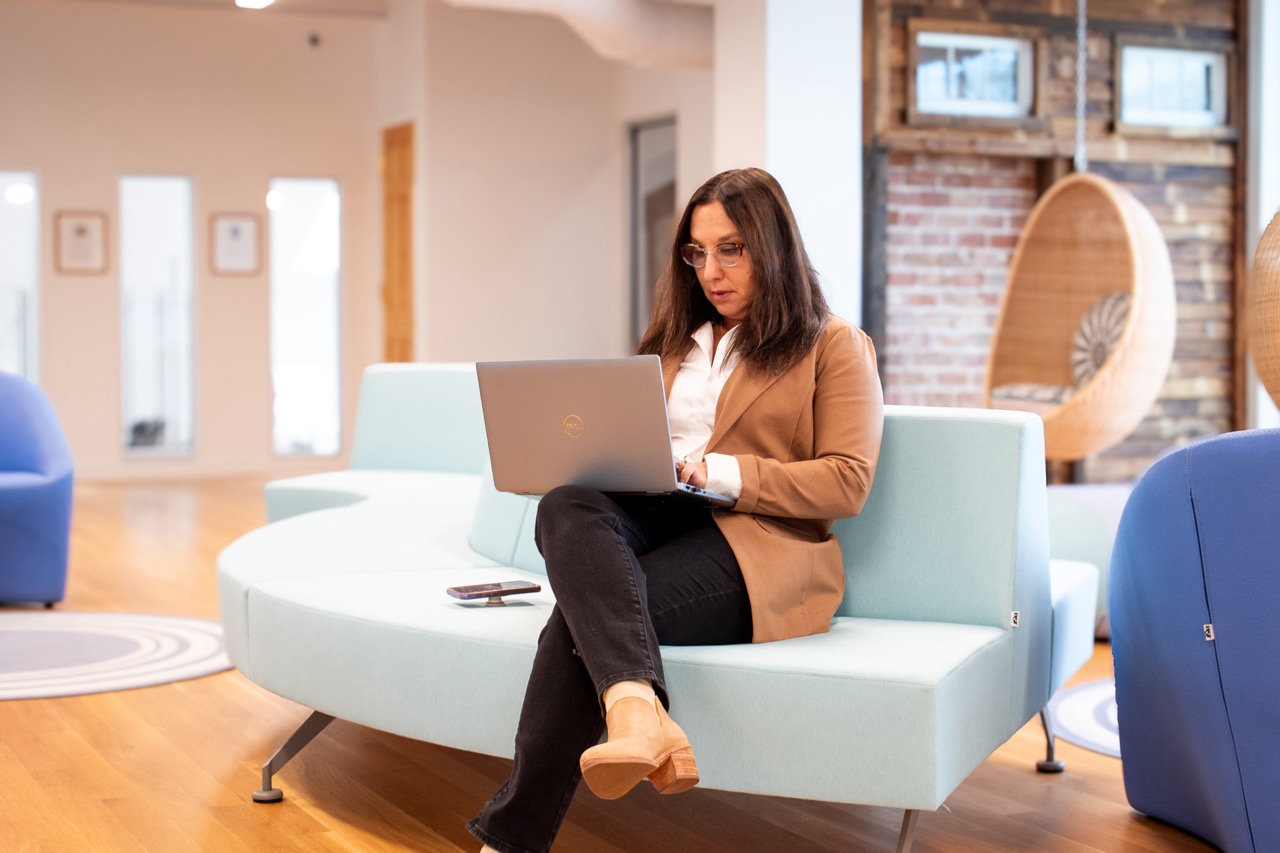 A woman works on a laptop while sitting on a modern couch in a bright lounge area.